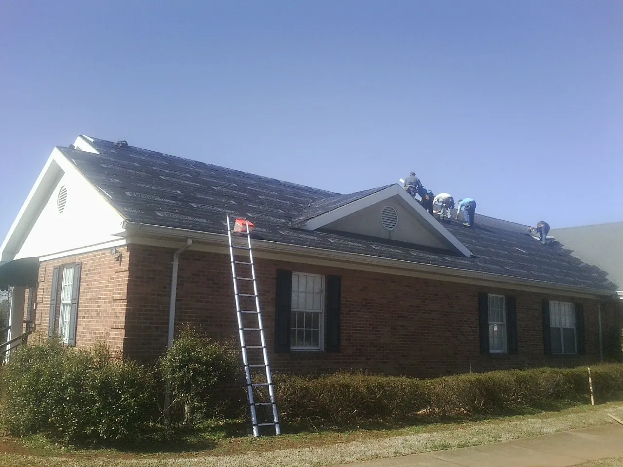 Crew tearing shingles off a brick ranch roof with a ladder set — Emergency Roof Repair work by our crew in Cement City