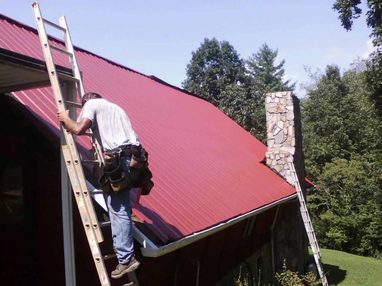 Roofer on a ladder beside a red metal roof and stone chimney — Soffit & Fascia Repair by Roofing Brothers in Cement City